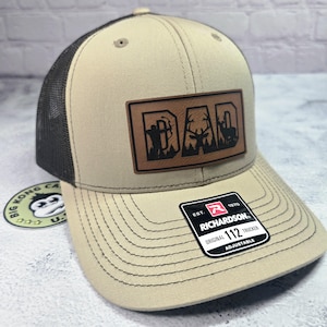 May include: Tan and black trucker hat with a leather patch that reads "DAD" with hunting-themed silhouettes. The hat features a mesh back, a curved brim, and an adjustable strap. The Richardson brand label is visible.