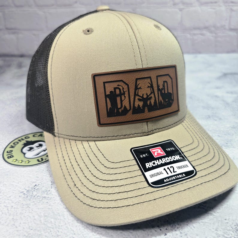 May include: Tan and black trucker hat with a leather patch that reads "DAD" with hunting-themed silhouettes. The hat features a mesh back, a curved brim, and an adjustable strap. The Richardson brand label is visible.