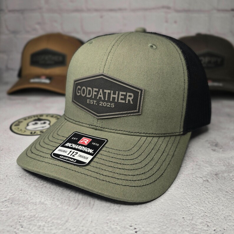 Puede incluir: Una gorra de camionero verde y negra con un parche negro que dice "GODFATHER EST. 2025". La gorra tiene una parte trasera de malla negra y una etiqueta Richardson 112.