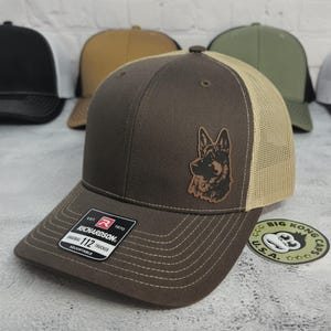 May include: Brown trucker hat with a tan mesh back and a German Shepherd patch. The hat has a curved brim and an adjustable strap. The Richardson brand label is visible on the front. The hat is part of a collection of hats.