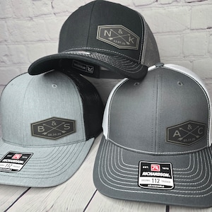 May include: Three black, gray, and white trucker hats with mesh back panels. Each hat features a different embroidered patch with a couple's initials and wedding date. The patches are in a brown, rectangular shape with a white background and black lettering. The hats are all adjustable.
