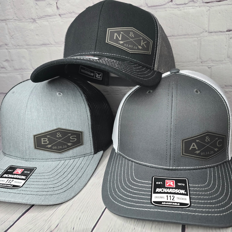 May include: Three black, gray, and white trucker hats with mesh back panels. Each hat features a different embroidered patch with a couple's initials and wedding date. The patches are in a brown, rectangular shape with a white background and black lettering. The hats are all adjustable.