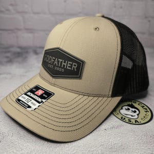Puede incluir: Una gorra de camionero beige y negra con un parche negro que dice "GODFATHER EST. 2025".