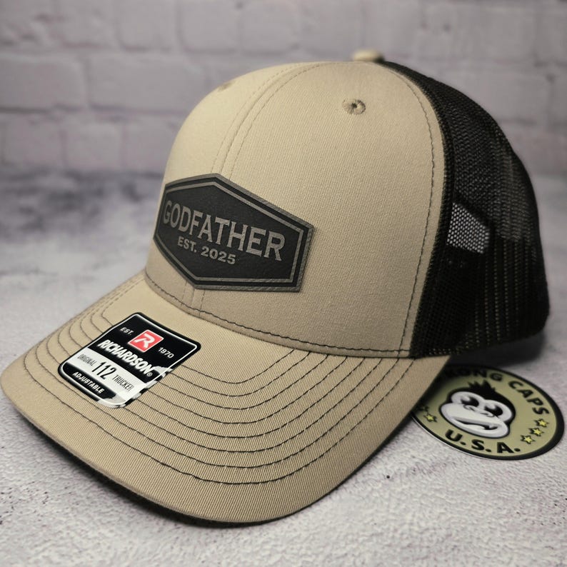 Puede incluir: Una gorra de camionero beige y negra con un parche negro que dice "GODFATHER EST. 2025".