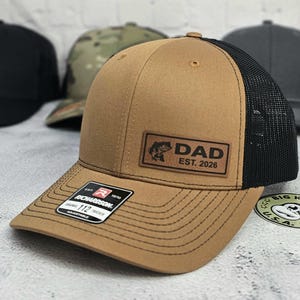 May include: A tan trucker hat with a black mesh back and a brown leather patch that reads "DAD EST. 2026" with a fish graphic. The hat has a curved brim and a Richardson 112 tag.