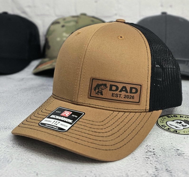 May include: A tan trucker hat with a black mesh back and a brown leather patch that reads "DAD EST. 2026" with a fish graphic. The hat has a curved brim and a Richardson 112 tag.
