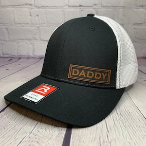 May include: Black and white mesh baseball cap with a brown leather patch that says "DADDY" on the front.