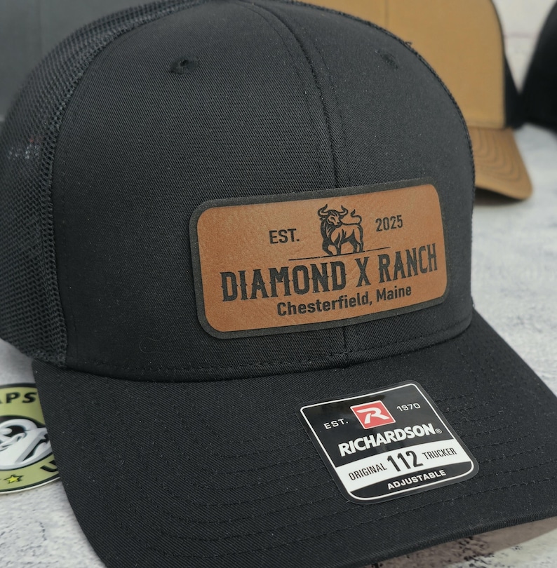 May include: Black trucker hat with a mesh back and a brown leather patch. The patch reads "DIAMOND X RANCH Chesterfield, Maine" with a bull graphic and the text "EST. 2025". The hat has a "RICHARDSON ORIGINAL 112 TRUCKER" tag.