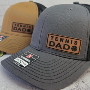 May include: Two baseball caps with the text "TENNIS DAD" on a leather patch. One cap is gray with a black mesh back, the other is tan with a black mesh back. Both have a Richardson 112 Trucker tag.