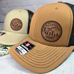 May include: Two trucker hats with a brown front panel and black mesh back. One hat has a tan front panel. Both hats feature a circular patch with a mountain and tree design. One patch reads "Yosemite National Park". The hats have a curved brim and adjustable strap.