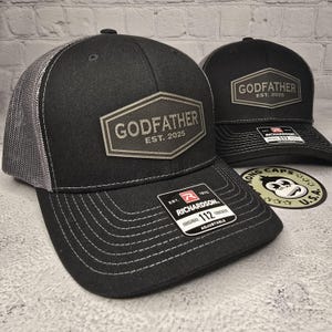 Puede incluir: Gorra de camionero negra y gris con un parche negro y blanco que dice "Godfather Est. 2025". La gorra es ajustable y tiene una parte trasera de malla.