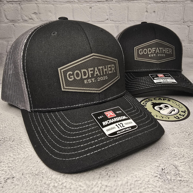 Puede incluir: Gorra de camionero negra y gris con un parche negro y blanco que dice "Godfather Est. 2025". La gorra es ajustable y tiene una parte trasera de malla.