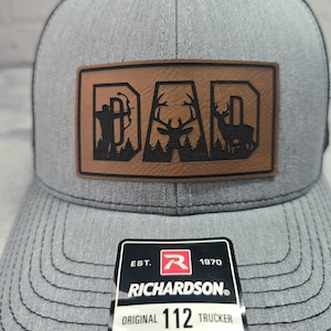 May include: Gray trucker hat with a brown leather patch that reads "DAD" with deer and archer silhouettes. The hat has a curved brim and a mesh back. The Richardson brand tag is visible.