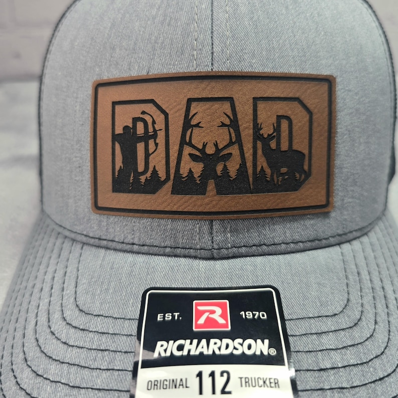 May include: Gray trucker hat with a brown leather patch that reads "DAD" with deer and archer silhouettes. The hat has a curved brim and a mesh back. The Richardson brand tag is visible.