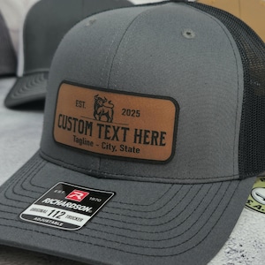 May include: A gray trucker hat with a black mesh back and a brown leather patch. The patch has the text "CUSTOM TEXT HERE" and the year "2025". The hat is adjustable and has the Richardson brand label.