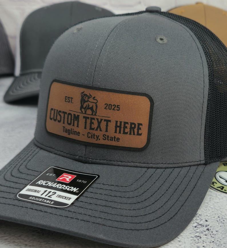 May include: A gray trucker hat with a black mesh back and a brown leather patch. The patch has the text "CUSTOM TEXT HERE" and the year "2025". The hat is adjustable and has the Richardson brand label.