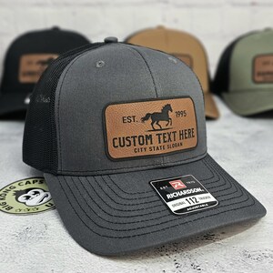 K&ouml;nnte beinhalten: Graue Trucker-Kappe mit schwarzem Mesh-R&uuml;cken und braunem Lederaufn&auml;her. Der Aufn&auml;her zeigt eine Pferdesilhouette, den Text "EST. 1995" und Platz f&uuml;r individuellen Text. Die Kappe ist verstellbar.
