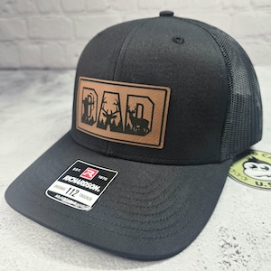 May include: Black Richardson 112 trucker hat with a brown leather patch. The patch features the word "DAD" with a forest and deer silhouette design. The hat has a mesh back and an adjustable strap.