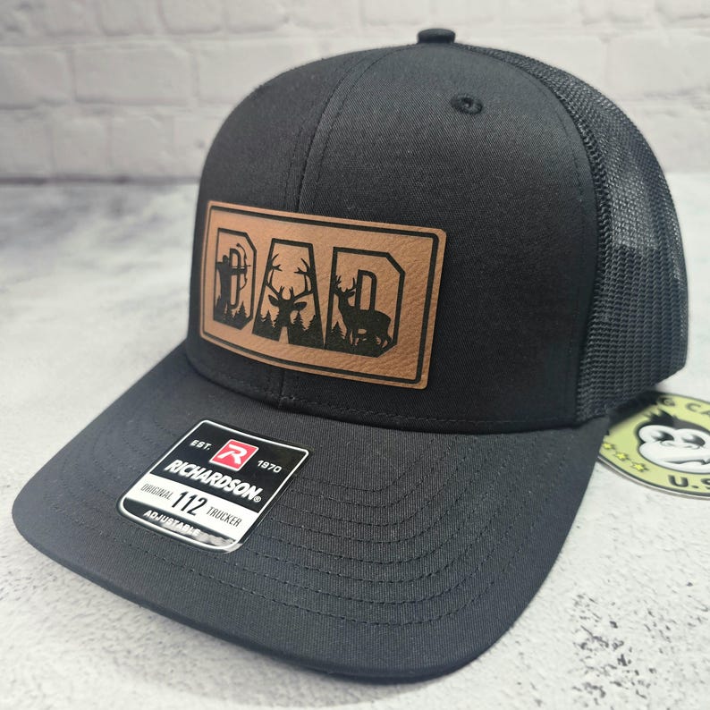 May include: Black Richardson 112 trucker hat with a brown leather patch. The patch features the word "DAD" with a forest and deer silhouette design. The hat has a mesh back and an adjustable strap.