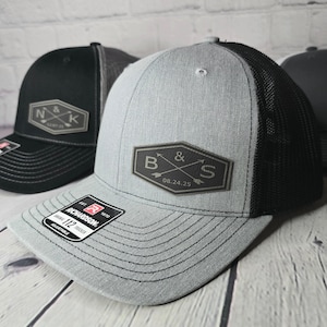 May include: A gray and black trucker hat with a black leather patch that says "B&S 08.24.25" in a vintage style font. The hat has a mesh back and a black adjustable strap.