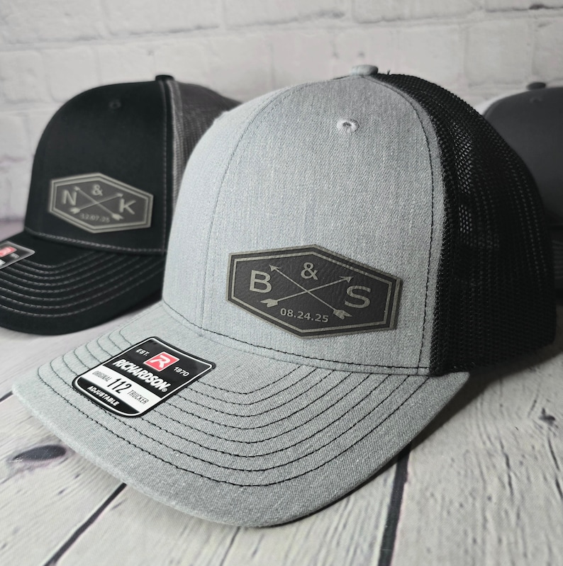 May include: A gray and black trucker hat with a black leather patch that says "B&S 08.24.25" in a vintage style font. The hat has a mesh back and a black adjustable strap.
