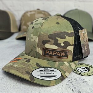 May include: Camouflage pattern snapback hat with a black mesh back. The hat features a brown leather patch with the word "PAPAW" and a "Multicam" tag. The brim has an orange stripe. The hat is from the brand "Yupoong".