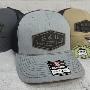May include: Gray trucker hat with a black mesh back and a leather patch. The patch features the letters "S&R" with the date "11.7.2020" and deer antlers. The hat has a curved brim and an adjustable strap.