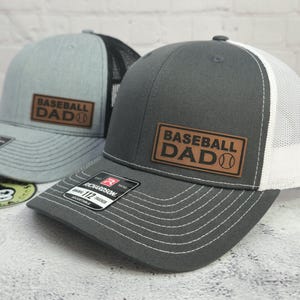 May include: Two baseball caps with the text "BASEBALL DAD" on a leather patch. One cap is gray with a black mesh back, and the other is dark gray with a white mesh back. Both have a curved brim and adjustable strap.