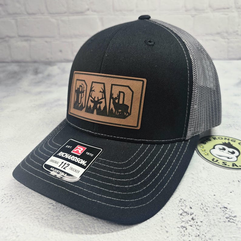 May include: Black and gray trucker hat with a brown leather patch that reads "DAD" with a deer silhouette design. The hat has a curved brim and an adjustable strap. The brand is Richardson.