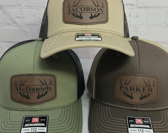 May include: Three Richardson 112 trucker hats with personalized leather patches. The hats are tan, olive green, and brown, each with a different name: Jacobson, McDaniels, and Parker. The hats have a mesh back and adjustable strap.