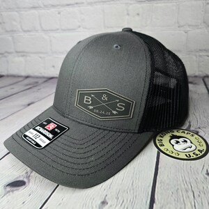 May include: A gray and black trucker hat with a black mesh back. The hat has a patch on the front with the letters "B & S" and the date "08.24.25".