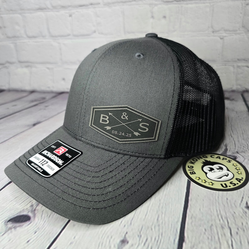 May include: A gray and black trucker hat with a black mesh back. The hat has a patch on the front with the letters "B & S" and the date "08.24.25".