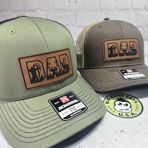 May include: Two trucker hats with leather patches that read "DAD" with hunting-themed graphics. One hat is olive green with a black mesh back, and the other is brown with a tan mesh back. Both have "RICHARDSON" tags.