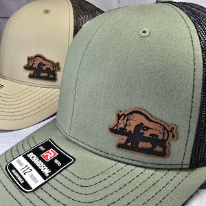 May include: Two trucker hats with a leather patch featuring a boar design. One hat is tan with a brown mesh back, the other is olive green with a black mesh back. Both have the Richardson 112 Trucker label.