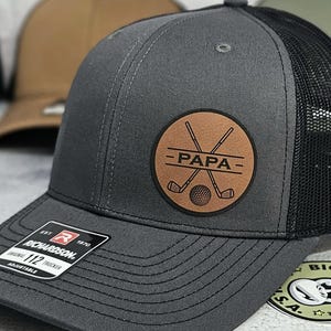 May include: Charcoal gray Richardson 112 trucker hat with a black mesh back. Features a brown leather patch with the word "PAPA" and crossed golf clubs and a golf ball. Adjustable fit.