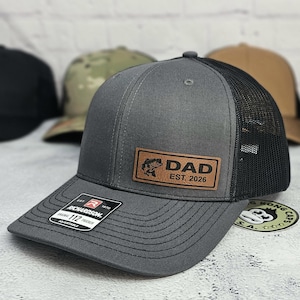 May include: A gray trucker hat with a black mesh back and a curved brim. The hat features a brown leather patch with the word "DAD" and a fish graphic, along with "EST. 2026". The hat has an adjustable strap.
