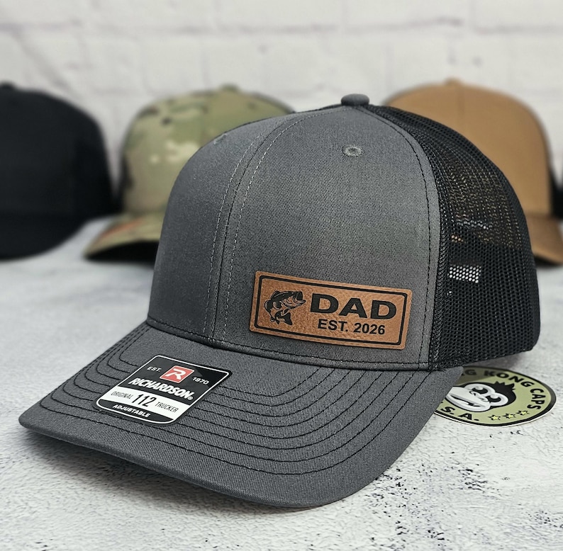 May include: A gray trucker hat with a black mesh back and a curved brim. The hat features a brown leather patch with the word "DAD" and a fish graphic, along with "EST. 2026". The hat has an adjustable strap.