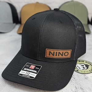 May include: Black Richardson 112 trucker hat with a mesh back and a curved brim. A brown leather patch with the name "NINO" is centered on the front. The hat has an adjustable strap and a "Big Kong Kaps" sticker.