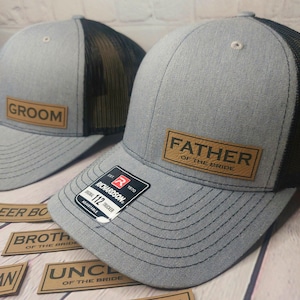 May include: Two gray and black trucker hats with brown leather patches. The patches say "Groom" and "Father of the Bride".