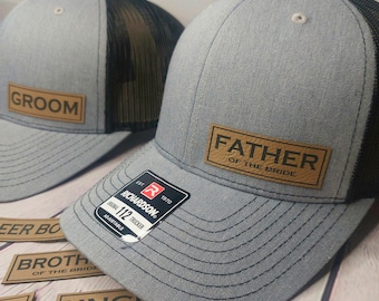 May include: Two gray and black trucker hats with brown leather patches. The patches say "Groom" and "Father of the Bride".