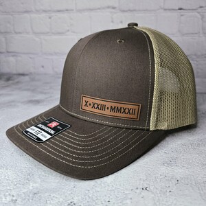 May include: A brown and beige trucker hat with a brown leather patch that says "X.XXIII.MMXXII".