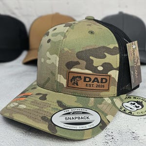 May include: Camouflage pattern baseball cap with a black mesh back. The cap features a leather patch with the word "DAD" and "EST. 2026" along with a fish graphic. The brim has an orange accent. The cap has a "SNAPBACK" closure.