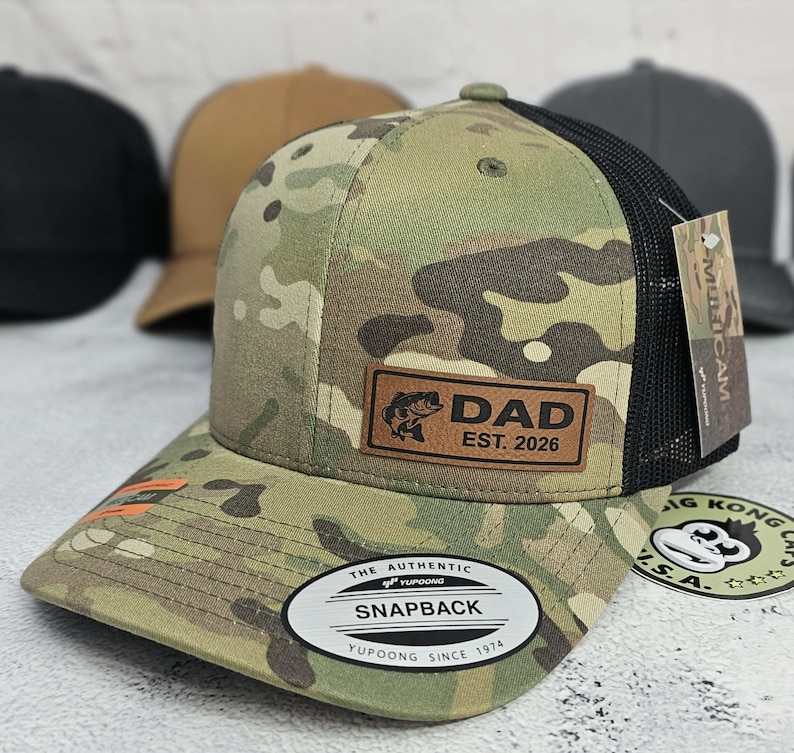 May include: Camouflage pattern baseball cap with a black mesh back. The cap features a leather patch with the word "DAD" and "EST. 2026" along with a fish graphic. The brim has an orange accent. The cap has a "SNAPBACK" closure.
