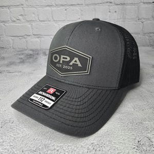 May include: A gray and black trucker hat with a silver patch that says "OPA EST. 2025". The hat has a black mesh back and a gray front.