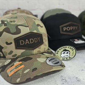May include: Camouflage baseball cap with the word "DADDY" on a brown patch. A black cap with "POPPY" on a brown patch is behind it. A "Big Kong Caps U.S.A." sticker is visible.