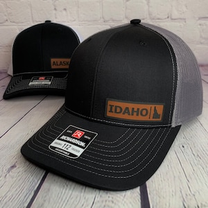 May include: Two black trucker hats with state patches. One hat features an "IDAHO" patch, and the other has an "ALASKA" patch. Both hats have a curved brim, a mesh back, and the "Richardson 112 Trucker" label.