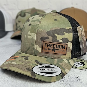May include: Camouflage pattern snapback hat with a black mesh back. The hat features a brown leather patch with the word "FREEDOM" and a rifle graphic. The hat also has a curved brim and a white tag that says "SNAPBACK".