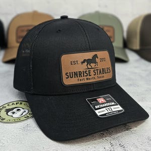 K&ouml;nnte beinhalten: Schwarze Trucker-Kappe mit Mesh-R&uuml;ckseite und gebogenem Schirm. Die Kappe hat einen braunen Lederaufn&auml;her mit dem Text "SUNRISE STABLES Fort Worth, Texas" und einer Pferdesilhouette. Die Kappe ist verstellbar und hat ein Richardson 112-Etikett.