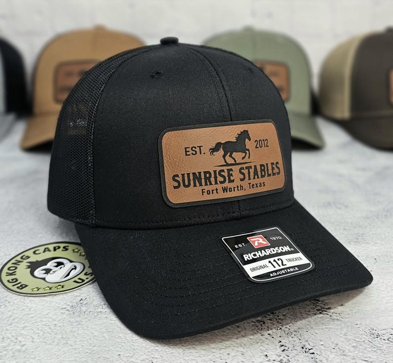 K&ouml;nnte beinhalten: Schwarze Trucker-Kappe mit Mesh-R&uuml;ckseite und gebogenem Schirm. Die Kappe hat einen braunen Lederaufn&auml;her mit dem Text "SUNRISE STABLES Fort Worth, Texas" und einer Pferdesilhouette. Die Kappe ist verstellbar und hat ein Richardson 112-Etikett.