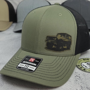 May include: Olive green trucker hat with a black mesh back and a patch featuring a vehicle graphic. The hat has a curved brim and an adjustable strap. The Richardson brand label is visible on the front.